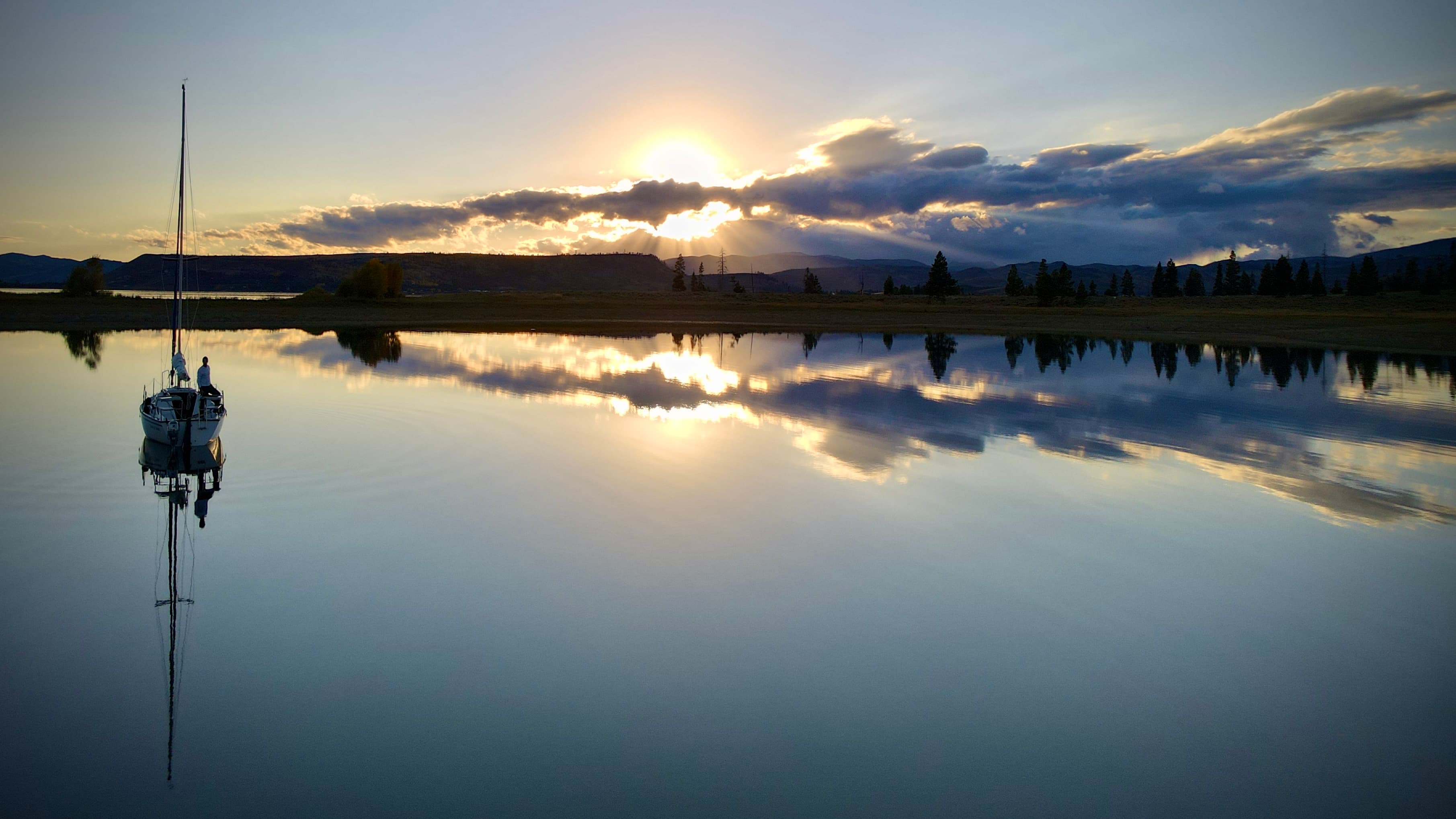 A sailboat at rest on perfectly glassy water at dusk. Crepuscular rays break through dramatic clouds. The boat and sky are reflected in the still surface below.