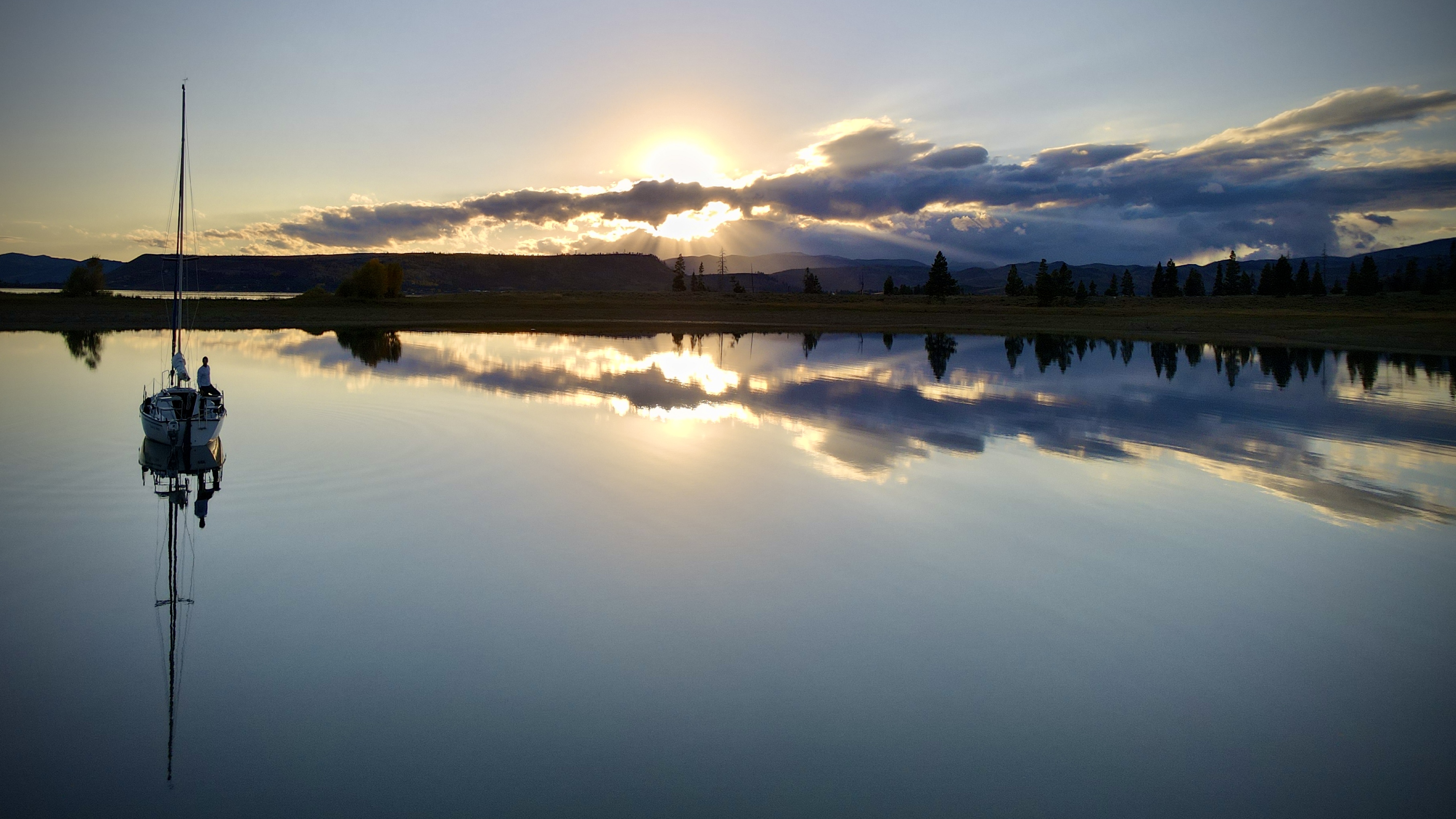 A sailboat at rest on perfectly glassy water at dusk. Crepuscular rays break through dramatic clouds. The boat and sky are reflected in the still surface below.
